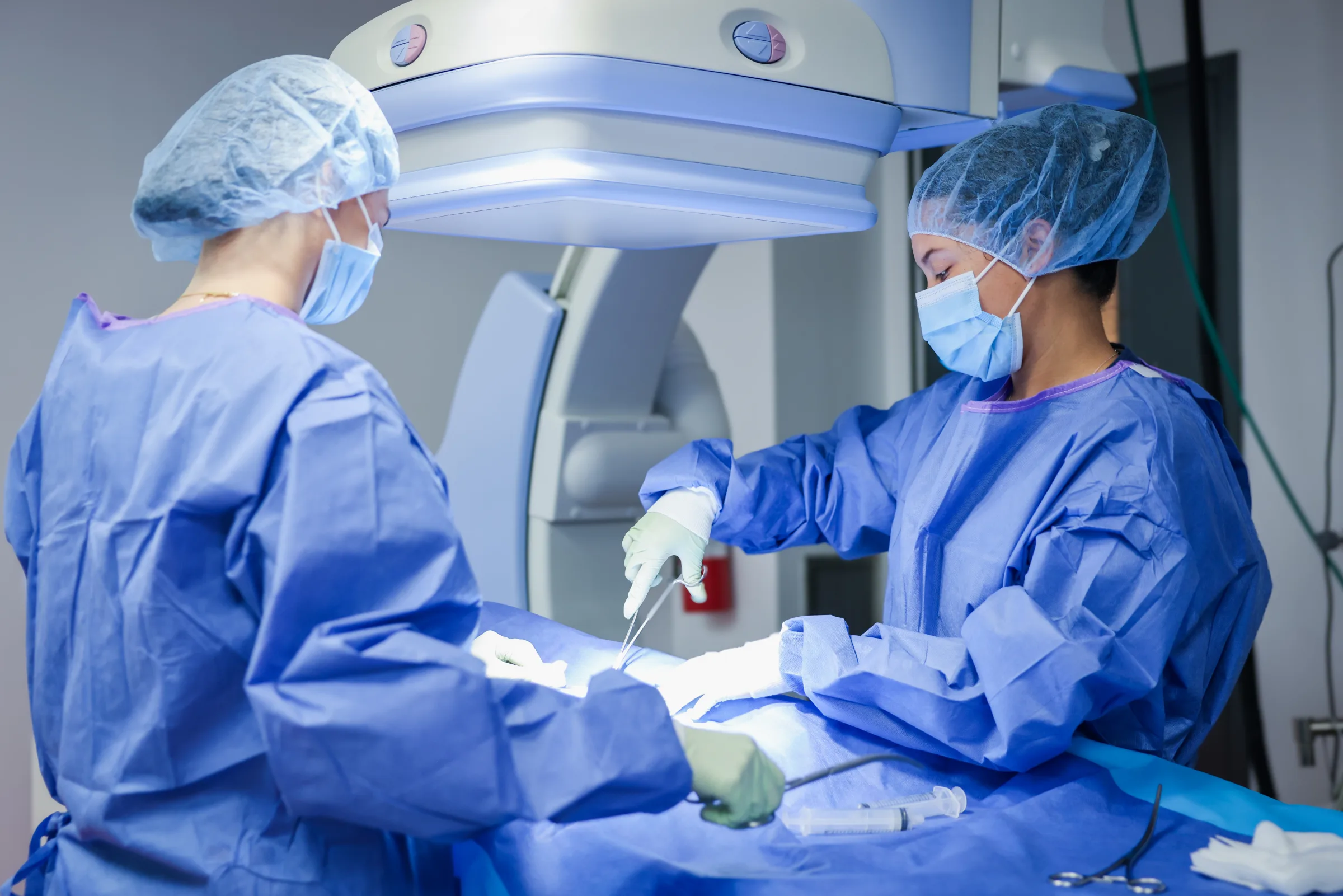 Two Synchrony Labs surgical staff in gowns and caps working the field under a C-arm in the procedure suite